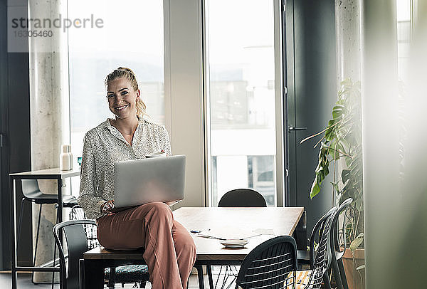 Lächelnde Geschäftsfrau sitzt mit Laptop auf einem Tisch im Büro