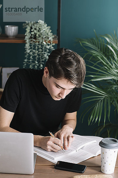 Junger männlicher Student  der in ein Buch schreibt  während er mit einem Laptop am Tisch sitzt und Hausaufgaben macht