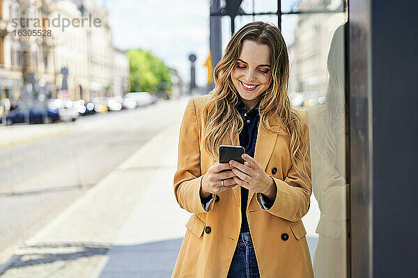 Lächelnde Frau  die beim Warten an der Straßenbahnhaltestelle eine Textnachricht über ihr Smartphone sendet