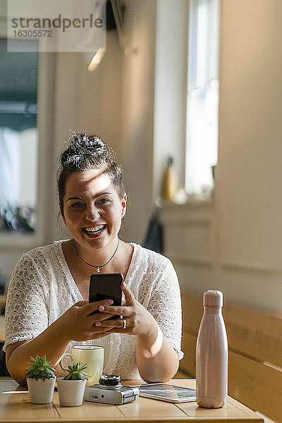 Fröhliche  üppige Frau  die ein Mobiltelefon benutzt  während sie am Tisch in einem Café sitzt