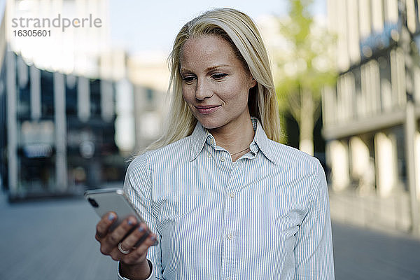 Lächelnde blonde Geschäftsfrau mit Smartphone im Finanzviertel