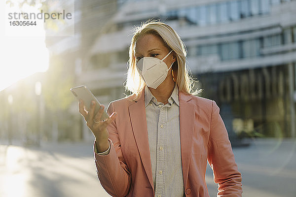 Geschäftsfrau mit Gesichtsmaske bei der Nutzung eines Smartphones im Finanzviertel während einer Pandemie