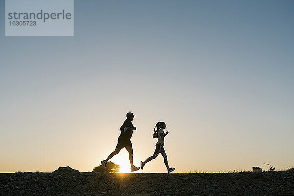 Silhouette eines Sportlers und einer Frau  die gegen den klaren Himmel bei Sonnenuntergang laufen