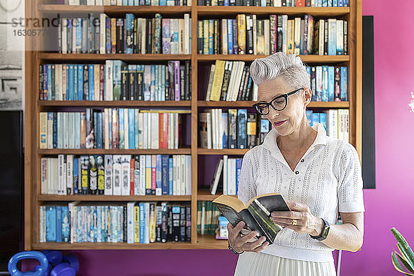 Aktive ältere Frau liest ein Buch  während sie zu Hause am Bücherregal steht