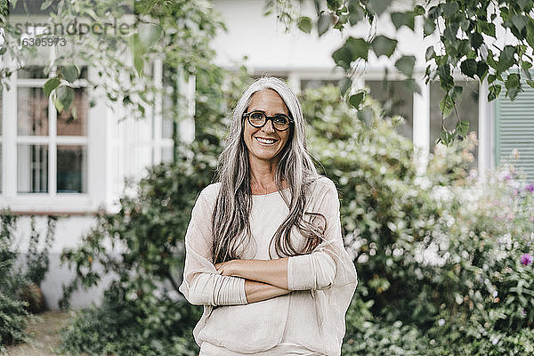 Porträt einer lächelnden Frau mit langen grauen Haaren und Brille  die im Garten steht