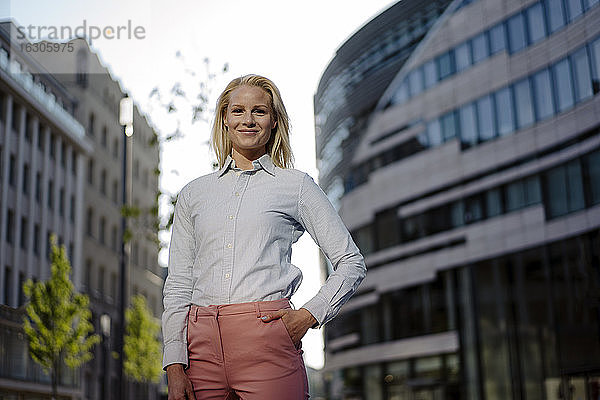 Lächelnde blonde Unternehmerin  die mit der Hand in der Tasche im Finanzviertel steht