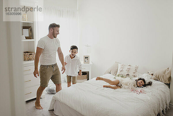 Verspielter lächelnder Vater  der seine Tochter über das Bett wirft  während der Sohn im Schlafzimmer steht