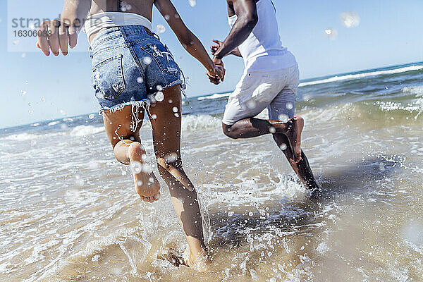 Verspieltes Paar hält sich an den Händen  während es im Meer gegen den klaren Himmel läuft
