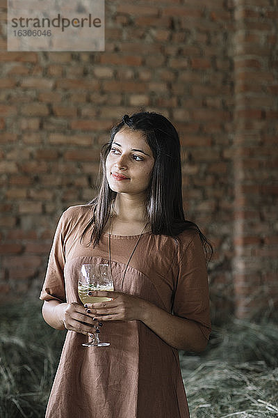 Nachdenkliche Frau hält einen frischen Cocktail in der Hand und blickt gegen eine Backsteinmauer an einer Scheune