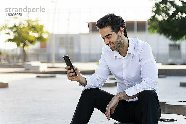 Gutaussehender Geschäftsmann  der ein Smartphone benutzt und gegen den Himmel in der Stadt sitzt