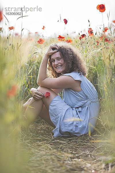 Lächelnde junge Frau mit Hand im Haar in einem Mohnfeld sitzend