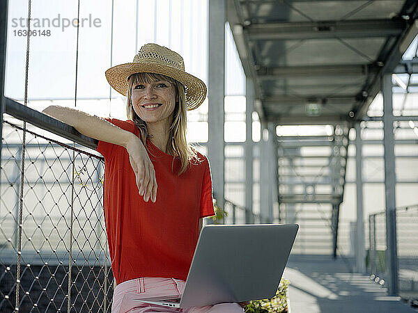 Lächelnde Geschäftsfrau mit Hut  die einen Laptop benutzt  während sie an einem Geländer im Gewächshaus sitzt