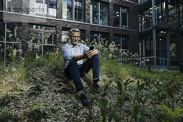 Geschäftsmann  der sein Smartphone benutzt  während er inmitten von Pflanzen vor einem Gebäude sitzt