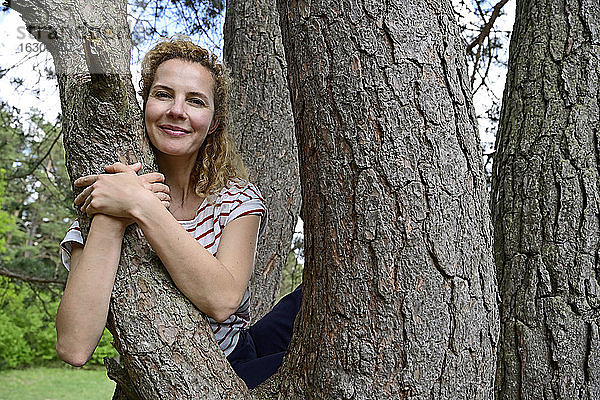 Lächelnde Frau  die sich umarmt  während sie auf einem Baum im Wald sitzt