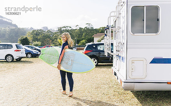 Frau  die an einem sonnigen Tag mit einem Surfbrett neben einem Wohnmobil spazieren geht