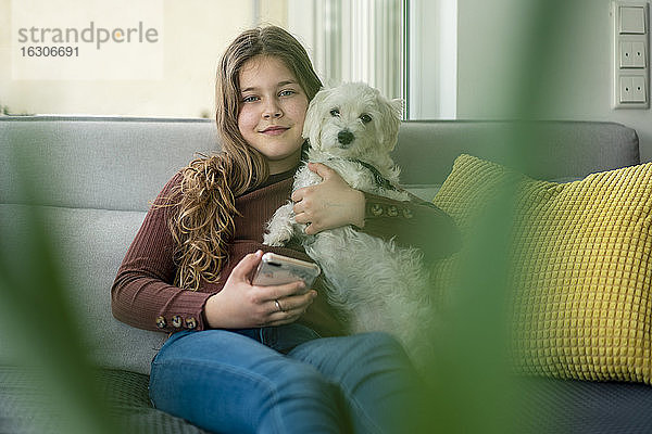 Lächelndes Mädchen  das ein Smartphone benutzt und einen Hund auf dem Sofa hält