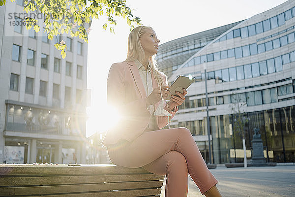 Von hinten beleuchtete Geschäftsfrau  die ein digitales Tablet hält  während sie auf einer Bank sitzt und in die Stadt blickt