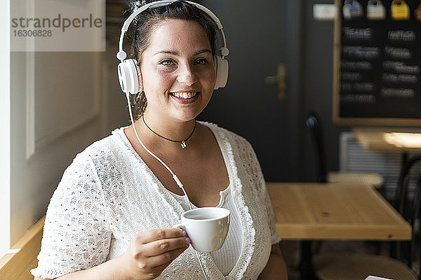 Nahaufnahme einer lächelnden jungen Frau  die eine Kaffeetasse hält und über Kopfhörer in einem Café Musik hört
