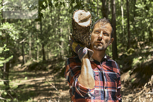 Nachdenklicher Holzfäller  der einen Baumstamm auf der Schulter trägt  während er im Wald in die Ferne schaut