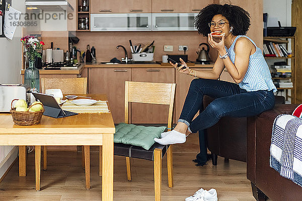 Unternehmerin mit lockigem Haar  die ihr Smartphone benutzt  während sie zu Hause Brot isst