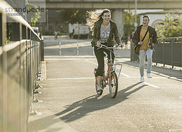 Freund läuft hinter einer glücklichen Freundin auf dem Fahrrad in der Stadt an einem sonnigen Tag