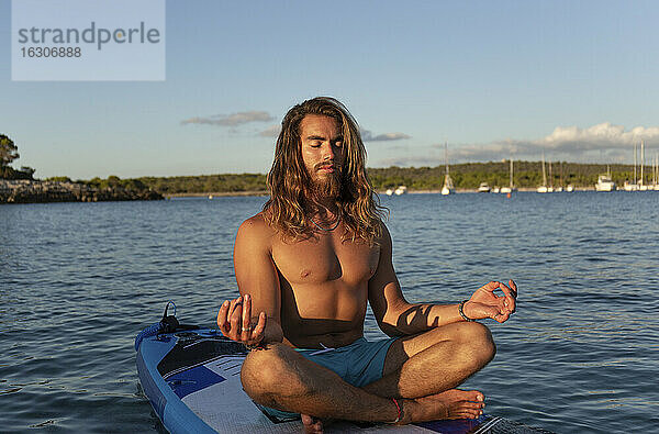 Junger Mann mit langen Haaren meditiert  während er auf einem Paddelbrett auf dem Meer gegen den Himmel bei Sonnenuntergang sitzt