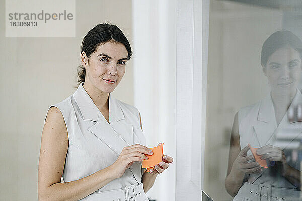 Frau hält Haftnotizen  während sie an einer Glaswand im Büro steht