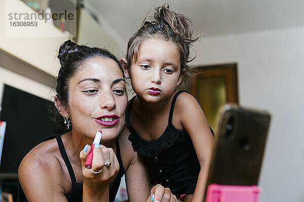 Mutter und Tochter tragen Lippenstift auf  während sie zu Hause mit dem Smartphone Videoaufnahmen machen