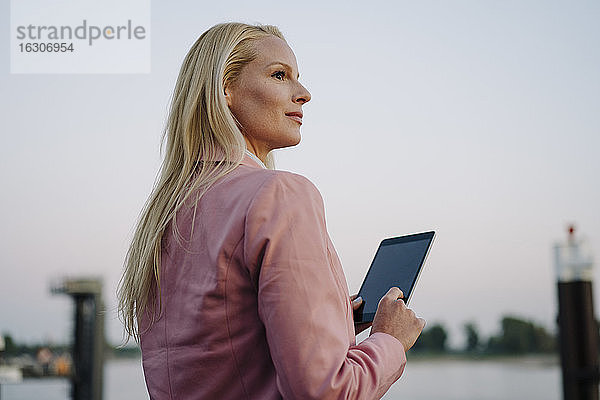 Nachdenkliche blonde Geschäftsfrau  die ein digitales Tablet hält und wegschaut