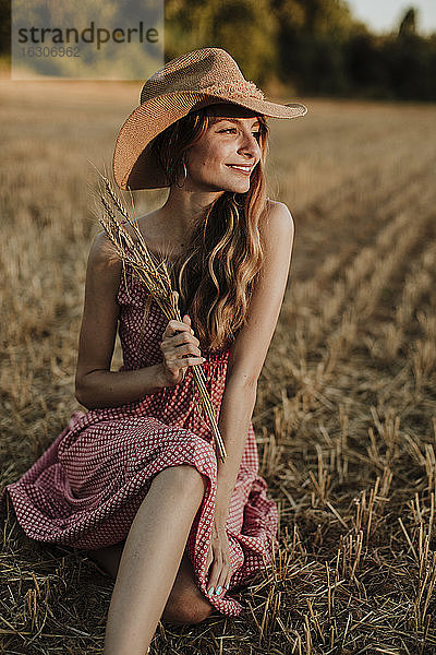 Glückliche Frau  die während des Sonnenuntergangs in einem Weizenfeld sitzt und wegschaut
