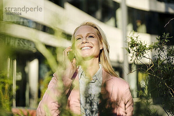 Lächelnde blonde Unternehmerin  die im Finanzdistrikt an einem sonnigen Tag mit einem Smartphone telefoniert
