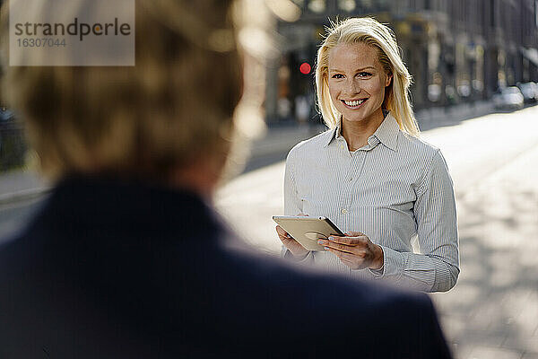 Lächelnde schöne weibliche Fachkraft  die ein digitales Tablet hält und einen Geschäftsmann in der Stadt betrachtet