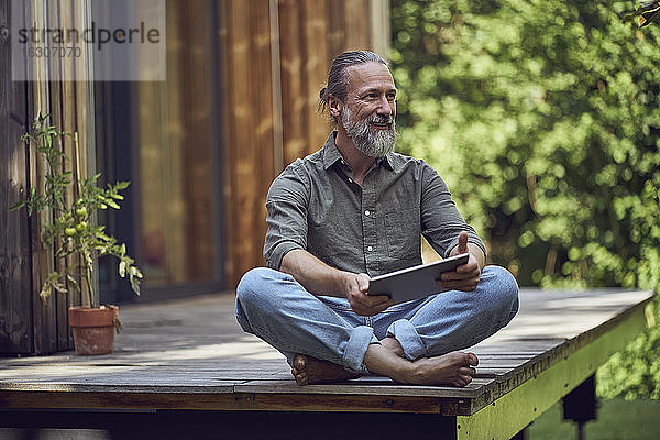 Bärtiger Mann mit digitalem Tablet  der vor einem kleinen Haus sitzt und nachdenkt