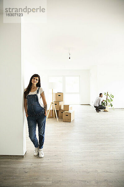 Schwangere Frau an der Wand stehend  während ihr Mann Monstera im neuen Haus arrangiert
