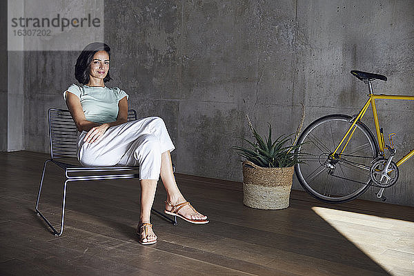 Lächelnde Geschäftsfrau entspannt sich auf einem Stuhl neben einer Pflanze und einem Fahrrad im Büro