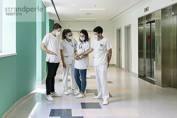 Ärzte  die chirurgische Masken tragen  diskutieren über ein digitales Tablet  während sie im Krankenhaus auf dem Flur stehen