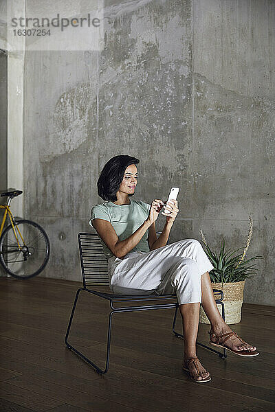 Geschäftsfrau  die ein Mobiltelefon benutzt  während sie auf einem Stuhl an der Wand im Büro sitzt