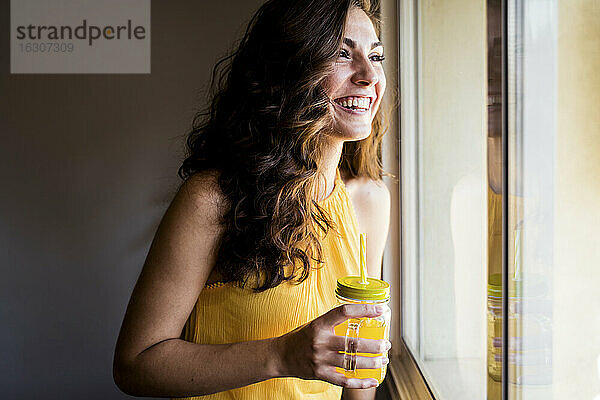 Glückliche Frau  die ein Einmachglas mit Saft in der Hand hält  während sie durch das Fenster zu Hause wegschaut