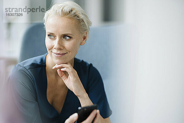 Nahaufnahme einer nachdenklichen Geschäftsfrau  die ein Smartphone hält  während sie im Büro sitzt