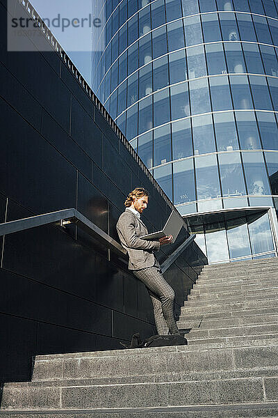 Geschäftsmann  der einen Laptop benutzt und sich auf einer Treppe im Stadtzentrum an die Wand lehnt  während die Sonne scheint