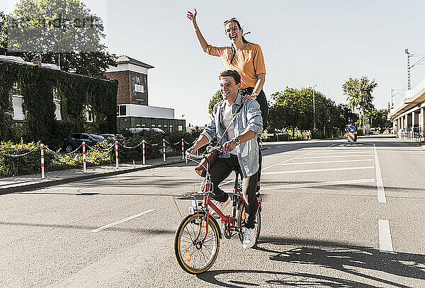 Fröhliche junge Frau steht hinter ihrem Freund  der auf der Straße Fahrrad fährt