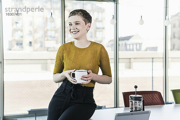 Lächelnde Geschäftsfrau sitzt auf einem Schreibtisch im Büro und hält einen Kaffeebecher