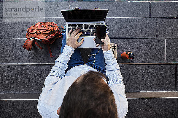 Männlicher Unternehmer  der auf einer Treppe sitzend ein Smartphone mit Laptop benutzt
