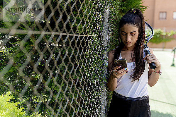 Eine Tennisspielerin benutzt ein Smartphone  während sie am Zaun des Tennisplatzes steht