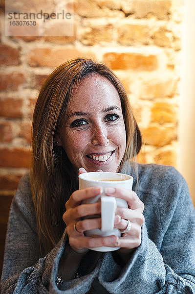 Frau lächelt  während sie eine Kaffeetasse gegen eine Backsteinmauer in einem Kaffeehaus hält