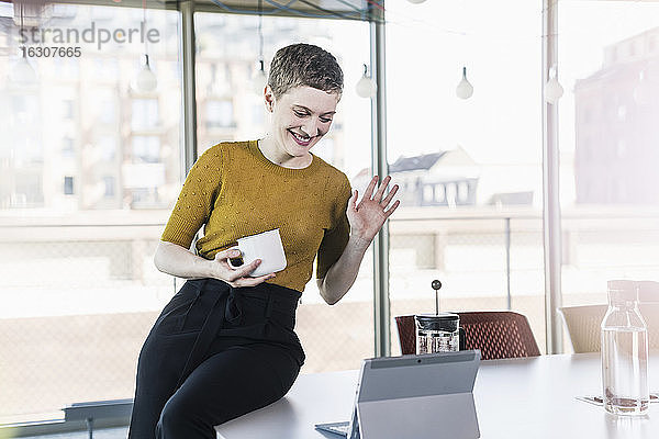 Lächelnde Geschäftsfrau sitzt auf einem Schreibtisch im Büro und führt eine Videokonferenz durch