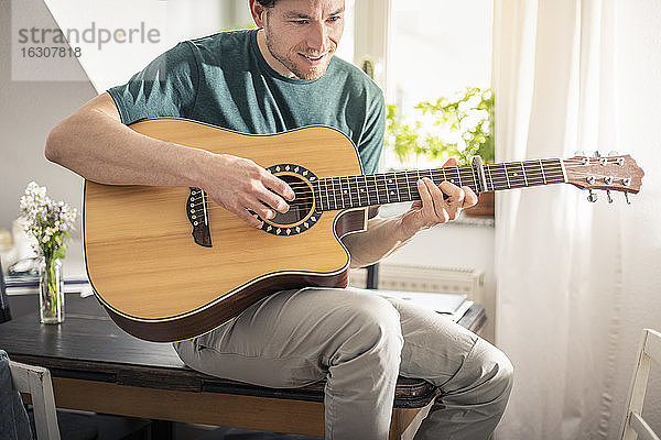 Lächelnder Mann spielt Gitarre  während er zu Hause auf dem Tisch sitzt