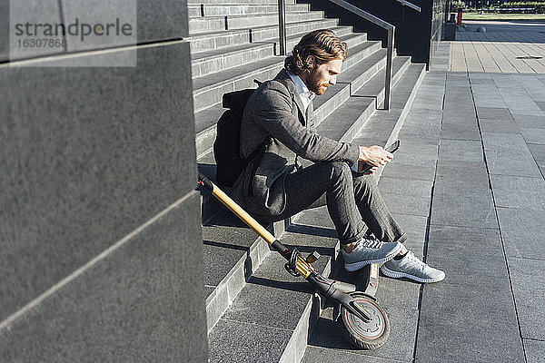 Männlicher Pendler  der ein digitales Tablet benutzt  während er mit einem Elektroroller auf einer Treppe im Stadtzentrum sitzt