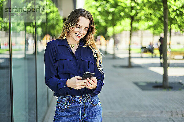 Lächelnde schöne Frau Text-Messaging durch Smartphone beim Stehen auf Gehweg in der Stadt