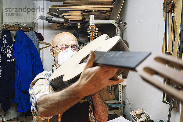 Handwerker mit Gesichtsmaske  der in der Werkstatt stehend eine Gitarre untersucht